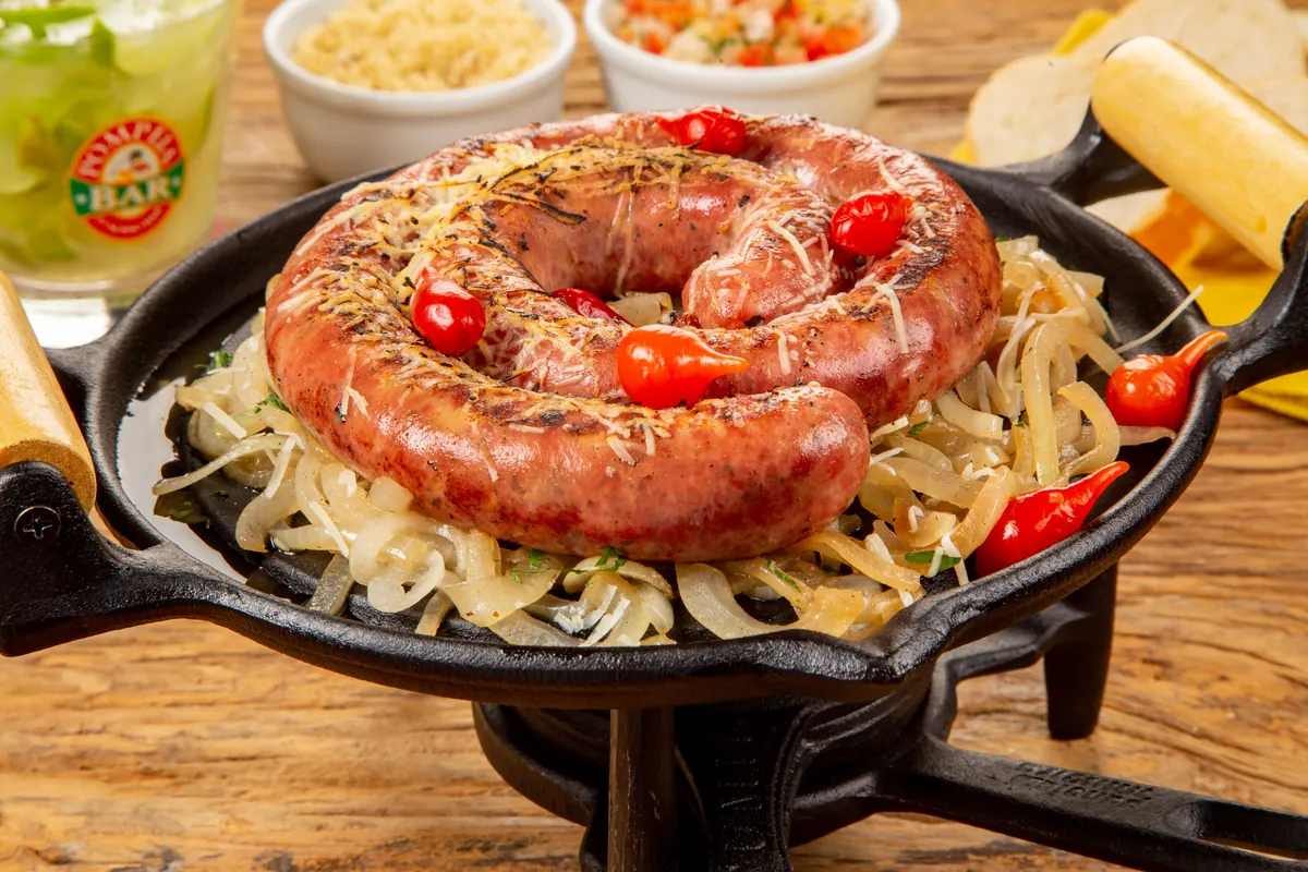 Calabresa artesanal de Bragança em espiral na chapa de ferro, com cebola, pimenta biquinho e drink ao fundo